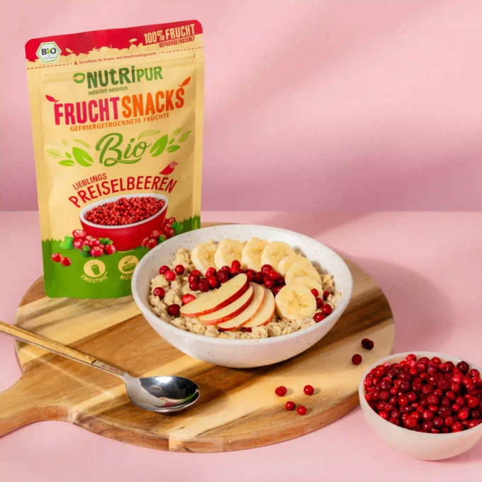 Hier ist ein Bild mit den gefriergetrockneten Preiselbeeren mit Müsli und Joghurt in einer Müslischale. Im Hintergrund ist die Verpackung abgebildet.