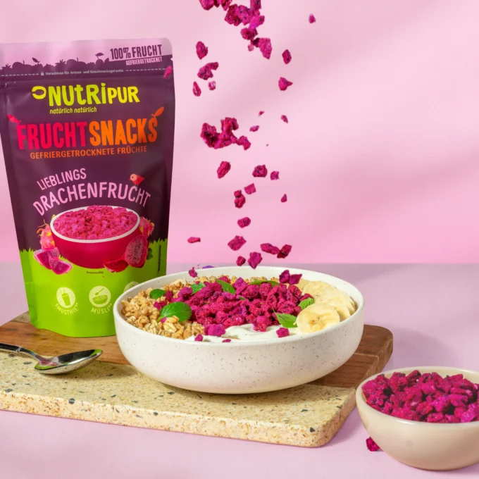 Hier ist ein Bild mit gefriergetrockneten Drachenfrucht als Müsli-Topping und Porridge-Topping in einer Schale. Im Hintergrund ist die Verpackung abgebildet.
