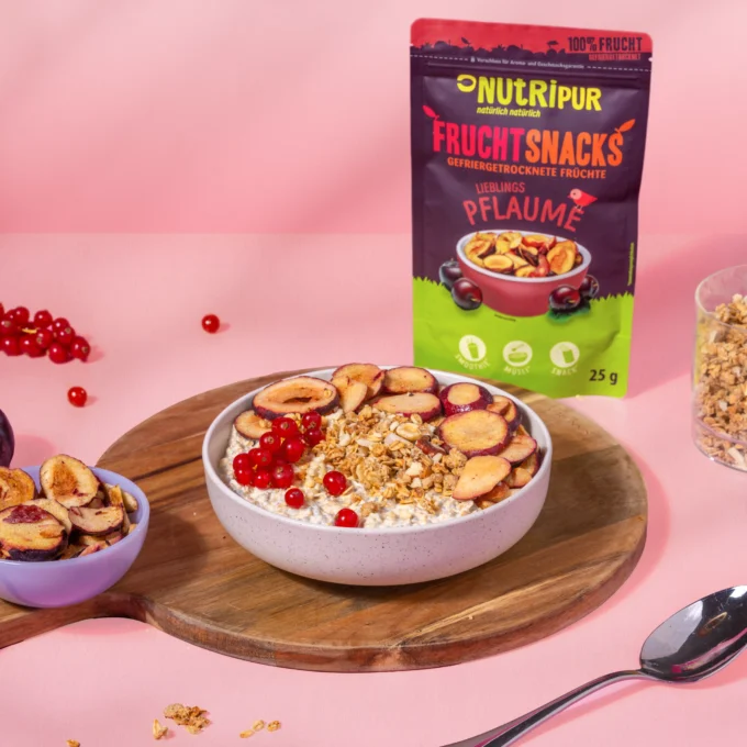 Hier ist ein Bild mit gefriergetrockneten Pflaumen als Müsli-Topping und Porridge-Topping in einer Schale. Im Hintergrund ist die Verpackung abgebildet.