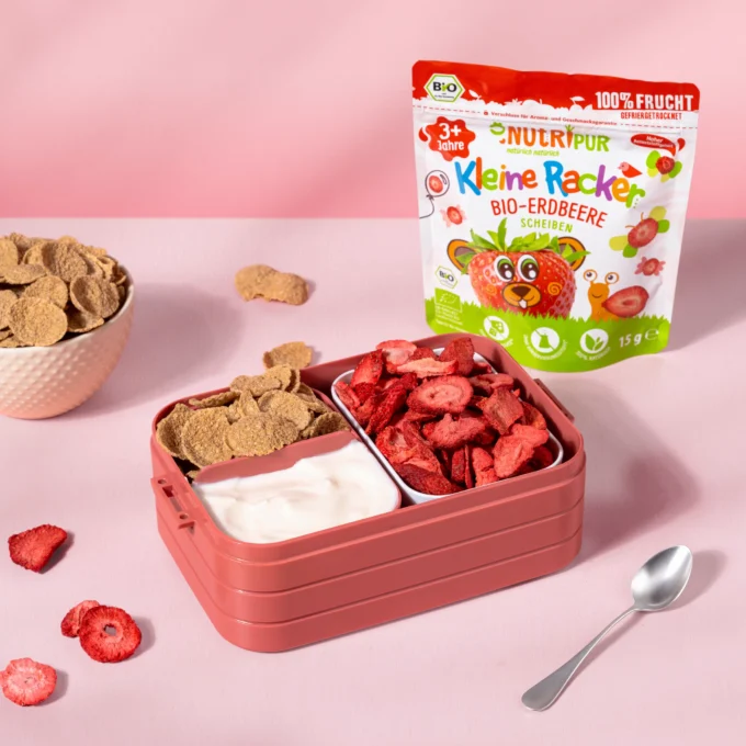 Hier ist ein Bild mit den kleinen Rackern der Sorte Bio-Erdbeere von Nutripur mit Joghurt und Flakes als Schulsnack in einer Brotdose . Im Hintergrund ist die Verpackung abgebildet.
