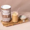 Das Bild zeigt eine Packung des Proteinpulvers der Sorte Super Isolat Whey Protein with chocolate taste. Zudem sieht man einen Smoothie, der mit dem Proteinpulver zubereitet wurde. Daneben steht eine Schale gefüllt mit dem Protein Pulver und einem Löffel. Alles ist hübsch auf einem Brettchen angerichtet.