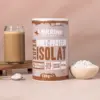 Das Bild zeigt eine Packung des Proteinpulvers der Sorte Super Isolat Whey Protein with chocolate taste. Zudem sieht man einen Smoothie, der mit dem Proteinpulver zubereitet wurde. Daneben steht eine Schale gefüllt mit dem Protein Pulver und einem Löffel. Alles ist hübsch auf einem Brettchen angerichtet.